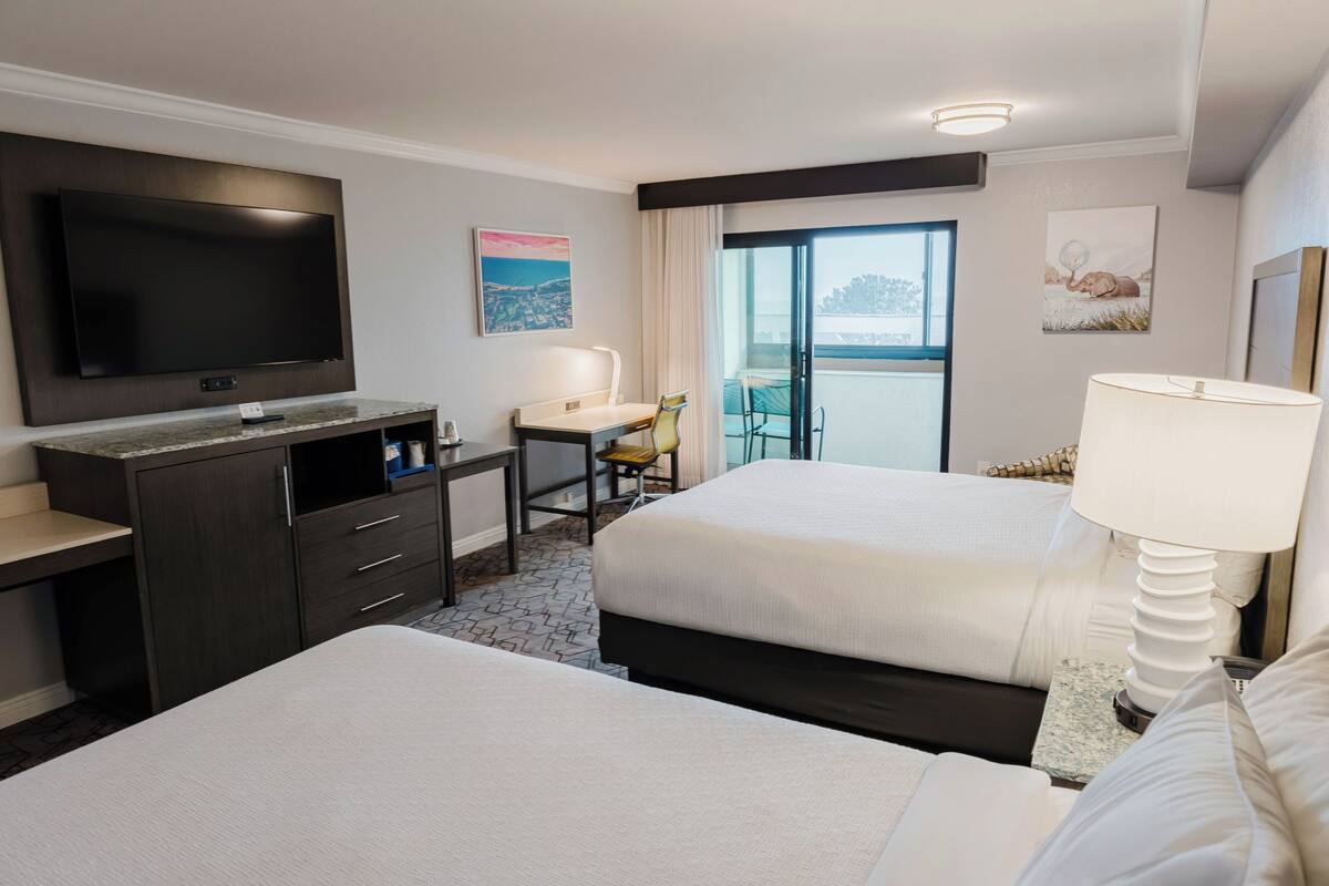 A hotel room with two beds, a desk, a TV, and a balcony with a view.