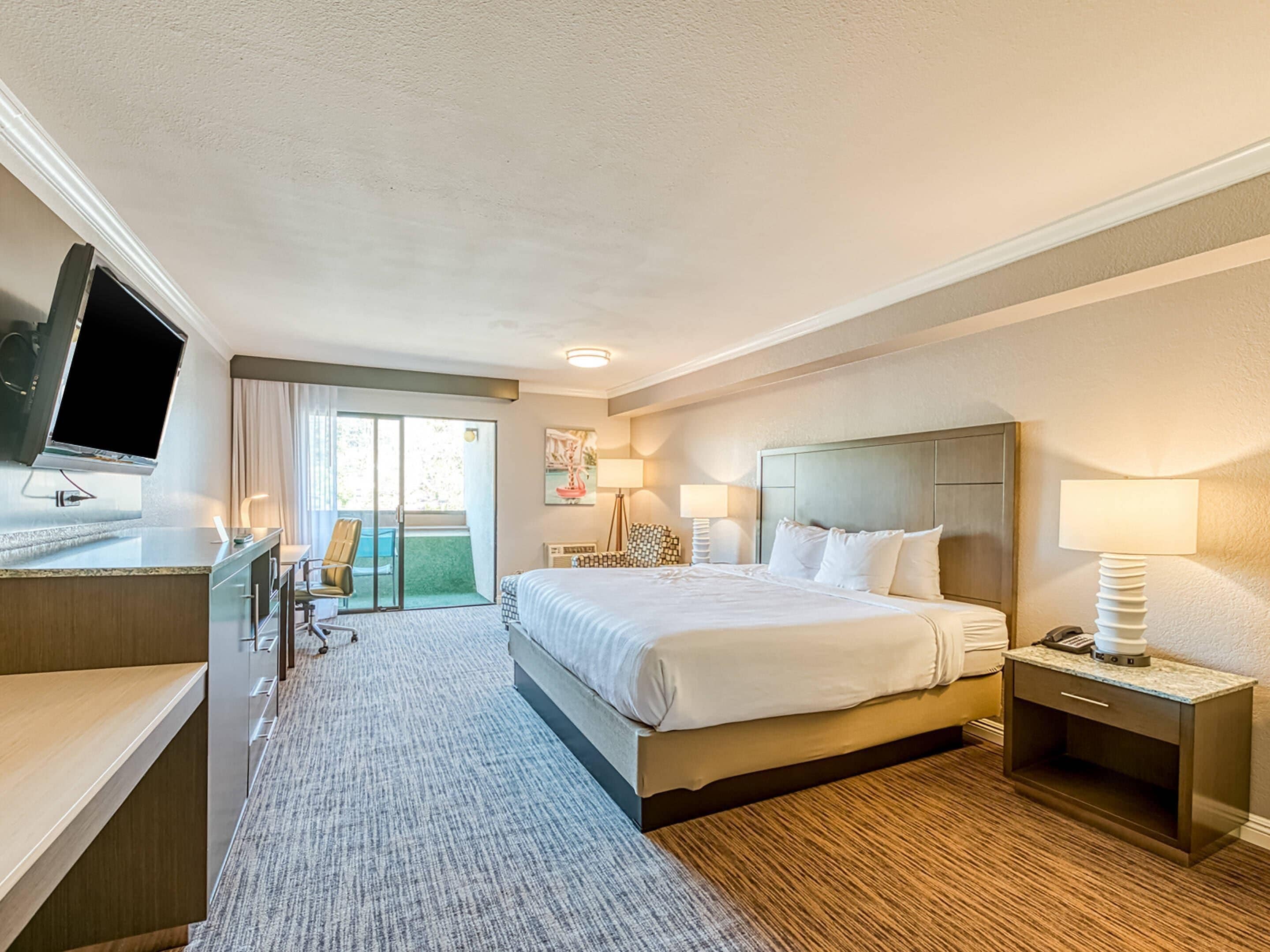 A hotel room with a bed, bedside tables, a desk with a flat-screen TV, a chair, and a view of the outdoors.