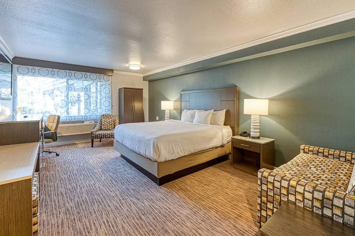 A hotel room with a large bed, two bedside tables with lamps, a desk, a chair, and a wardrobe.
