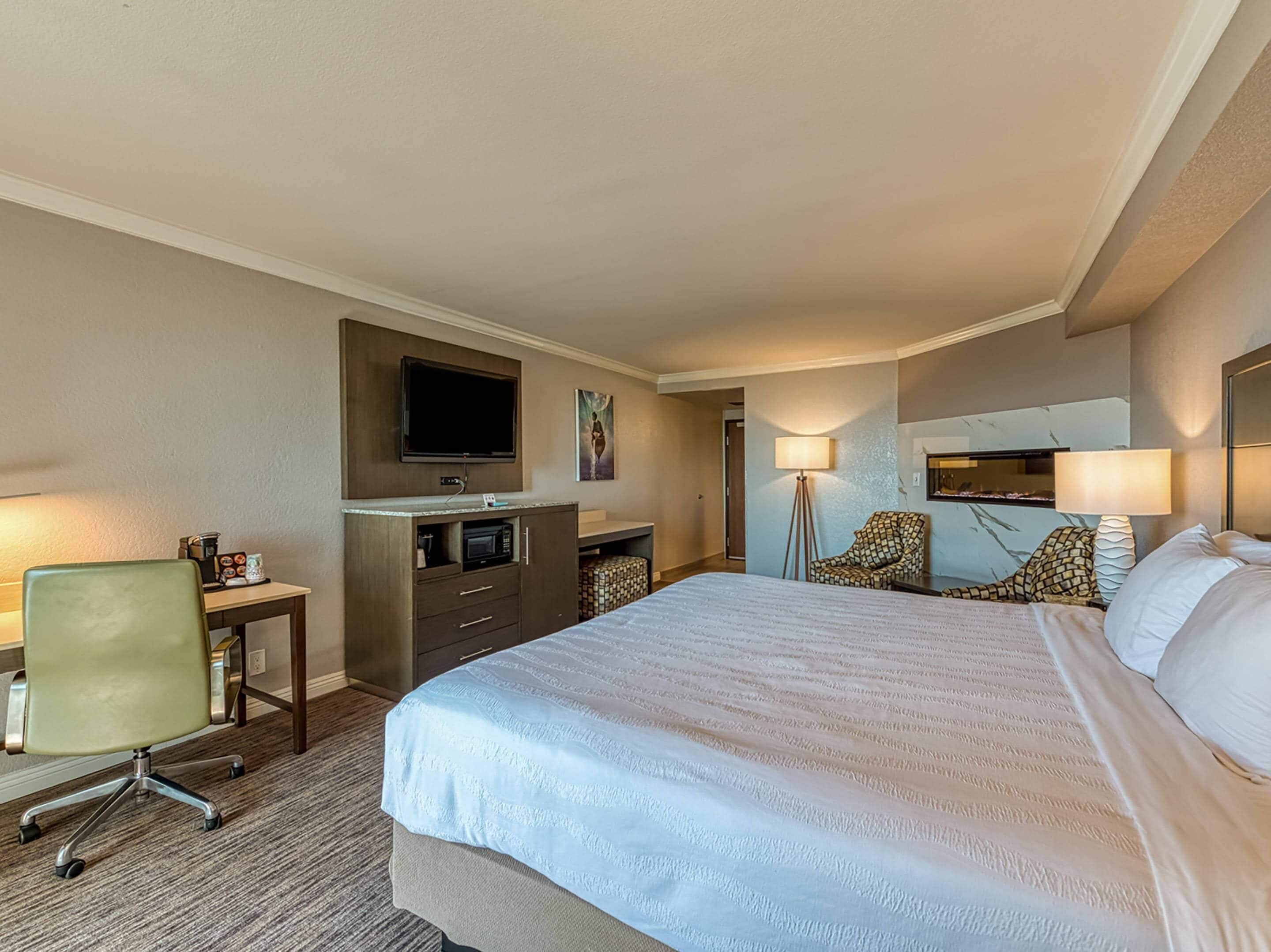 A hotel room with a large bed, a desk with a flat-screen TV, a chair, a lamp, and a small table with a coffee maker.