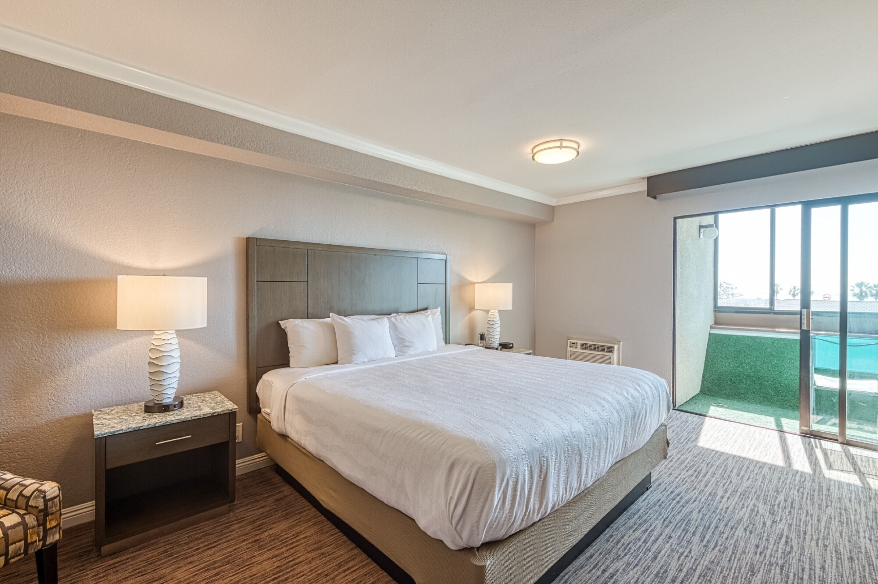 A hotel room with a large bed, two bedside tables with lamps, a headboard, and a view of a swimming pool through a sliding glass door.