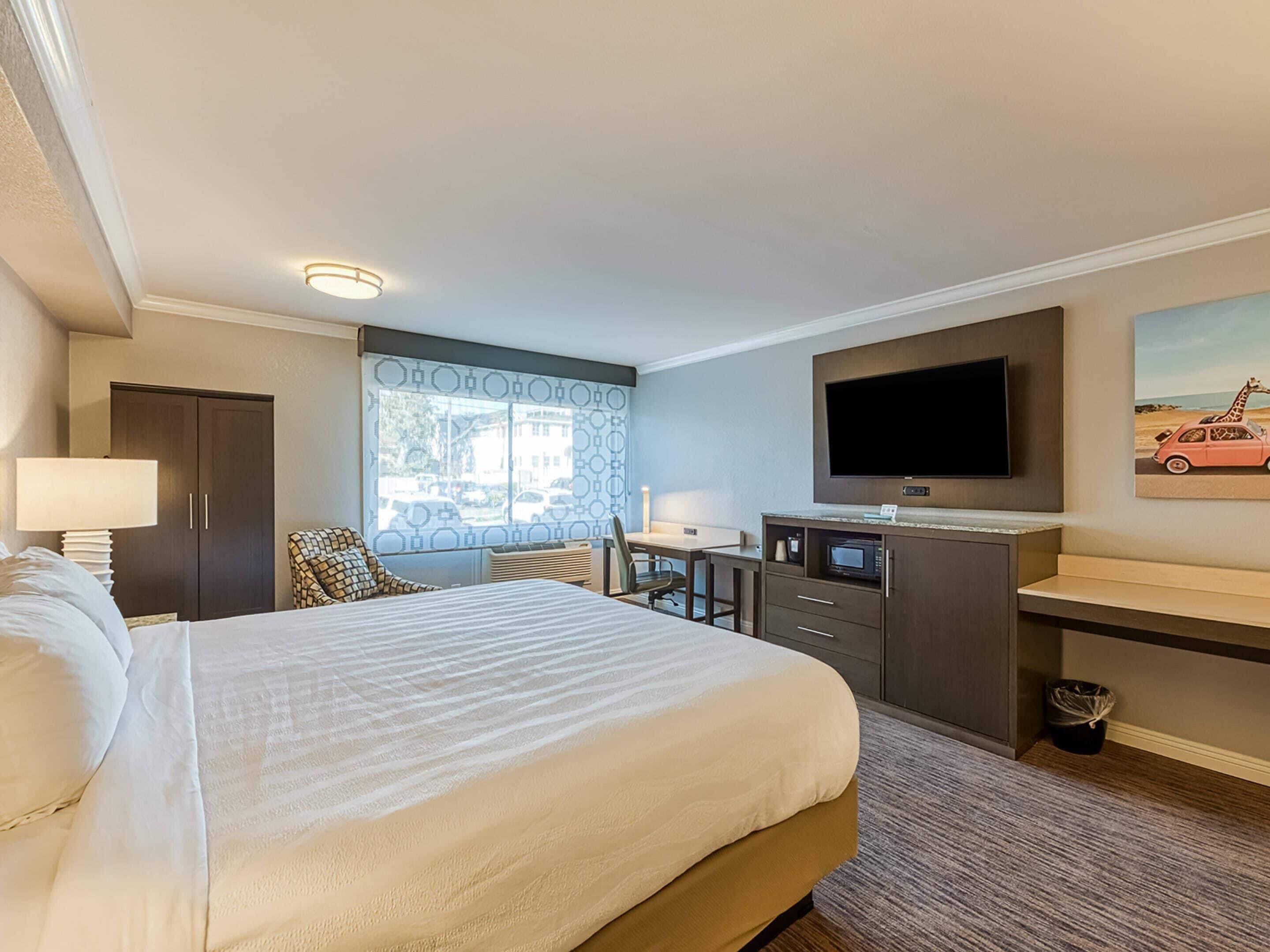 A hotel room with a large bed, a desk with a flat-screen TV, a window with a view of the street, and a door.