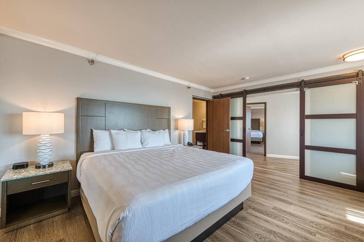 A hotel room with a large bed, two bedside tables with lamps, a wooden floor, and a sliding glass door leading to another room.