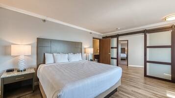 Una habitación de hotel con una cama grande, dos mesitas de noche con lámparas, suelo de madera y un armario corredero de vidrio que da a otra habitación.