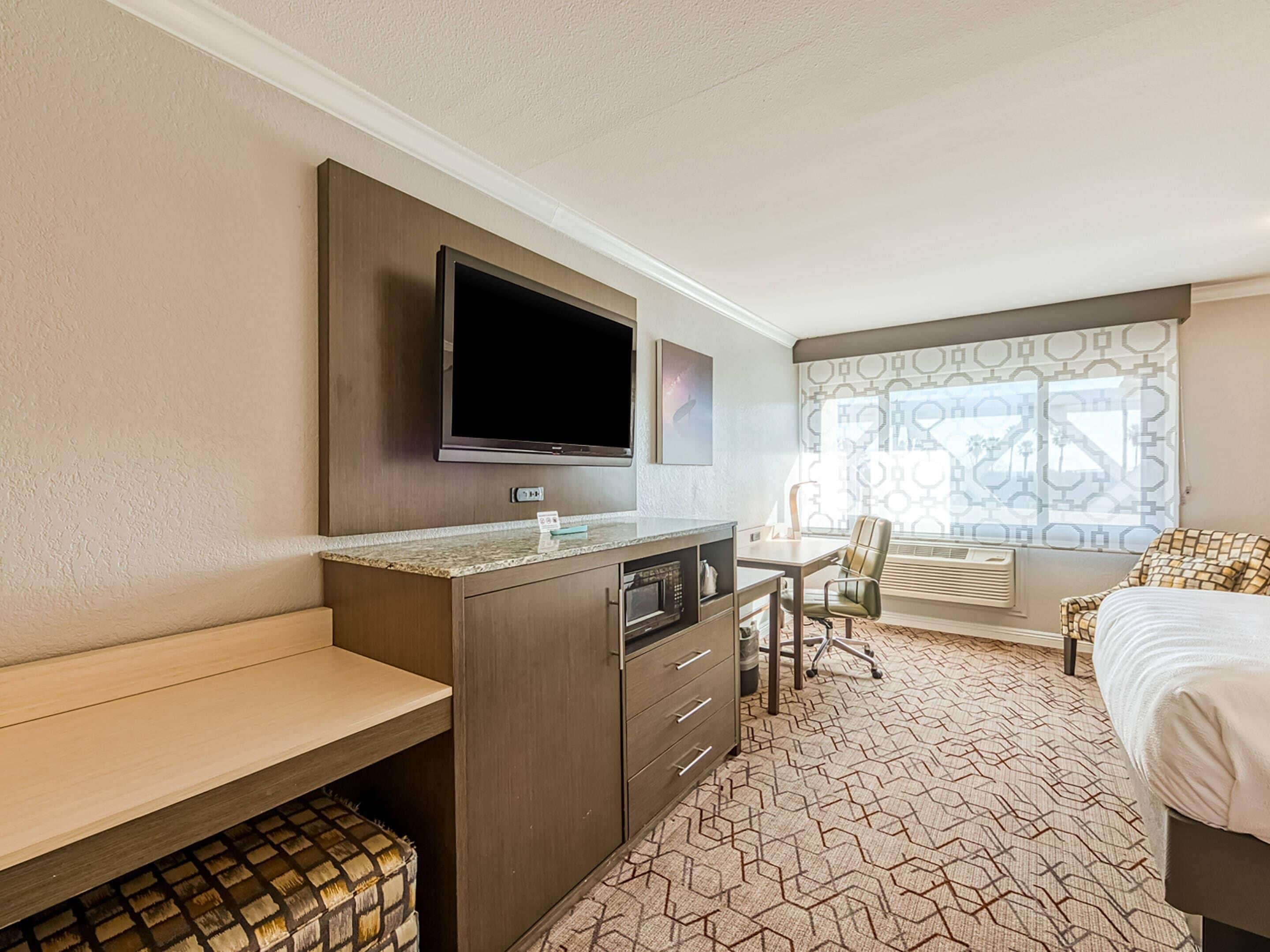 Quarto de hotel com cama, escrivaninha com cadeira, televisão e janela com cortinas estampadas.