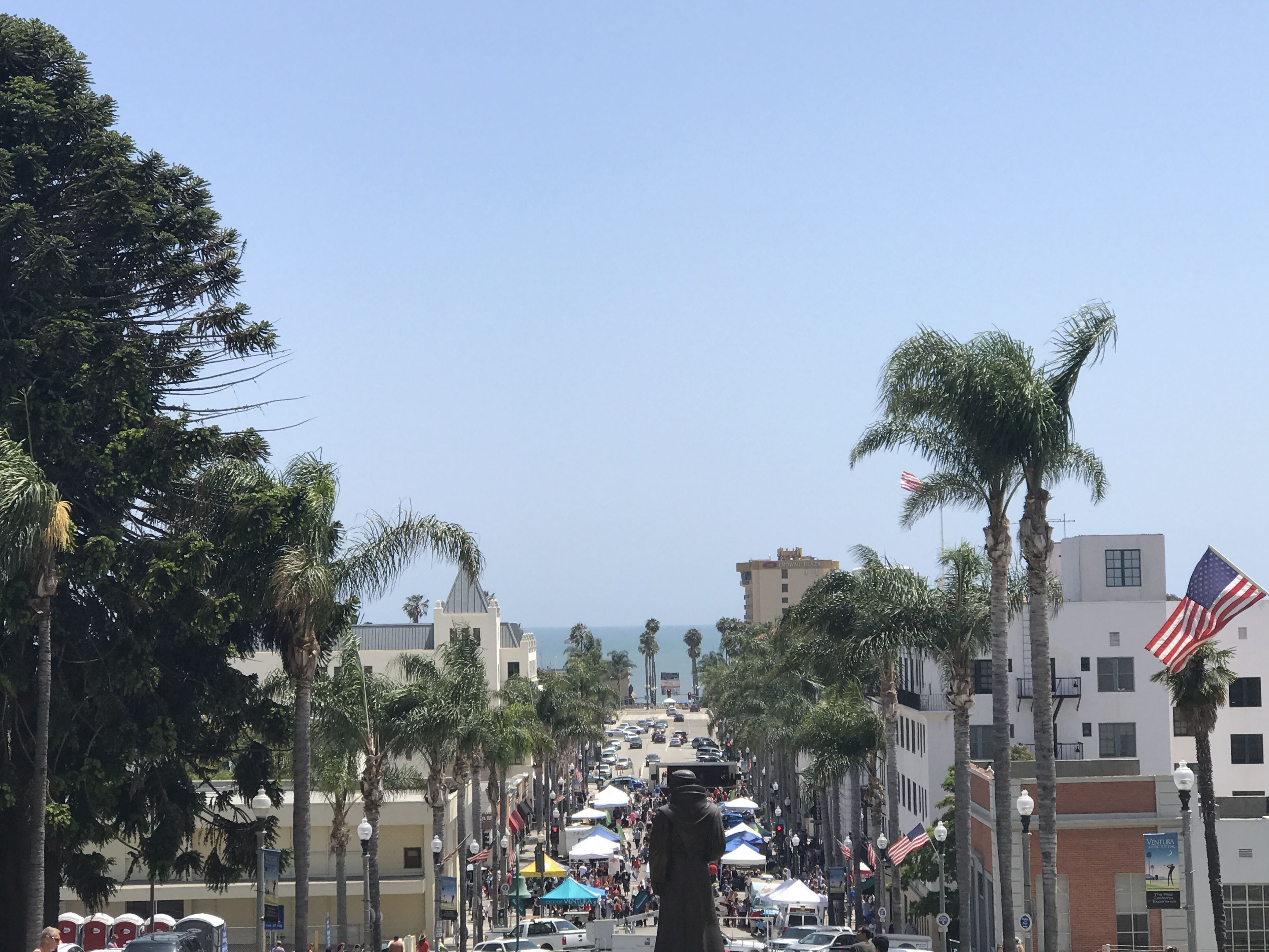 A street lined with palm trees and buildings, featuring a statue, American flags, and a clear sky.