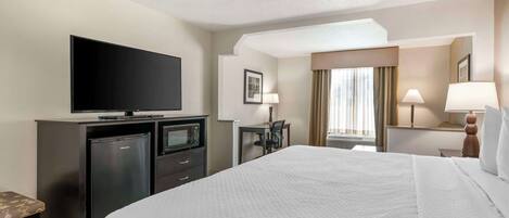 A hotel room with a large bed, a flat-screen TV, a minibar, a desk with a chair, and a window with curtains.