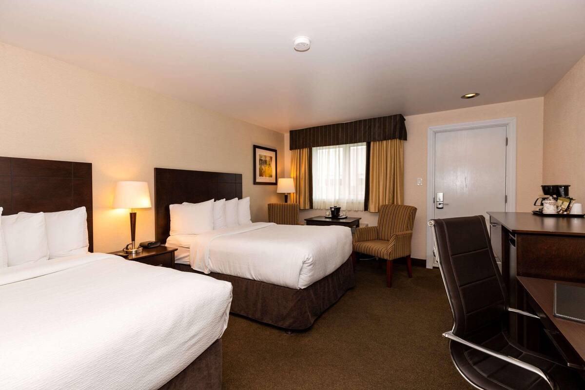 A hotel room with two beds, a desk, a chair, and a small table with a lamp.