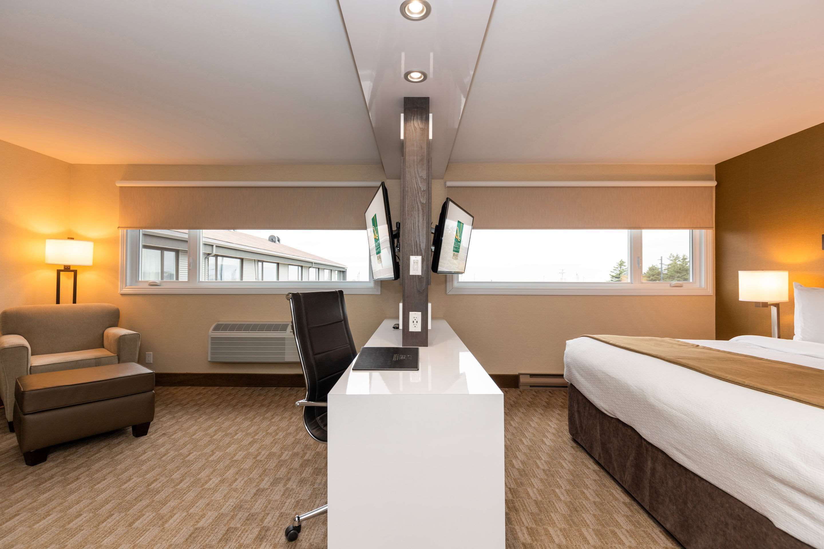 Ein Hotelzimmer mit einem Bett, einem Schreibtisch mit Computer, einem Stuhl, einer Lampe und einem Fenster mit Blick auf ein Gebäude.