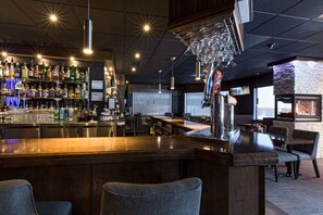 A bar area with a wooden counter, shelves of liquor bottles, hanging wine glasses, and a seating area with chairs and tables.