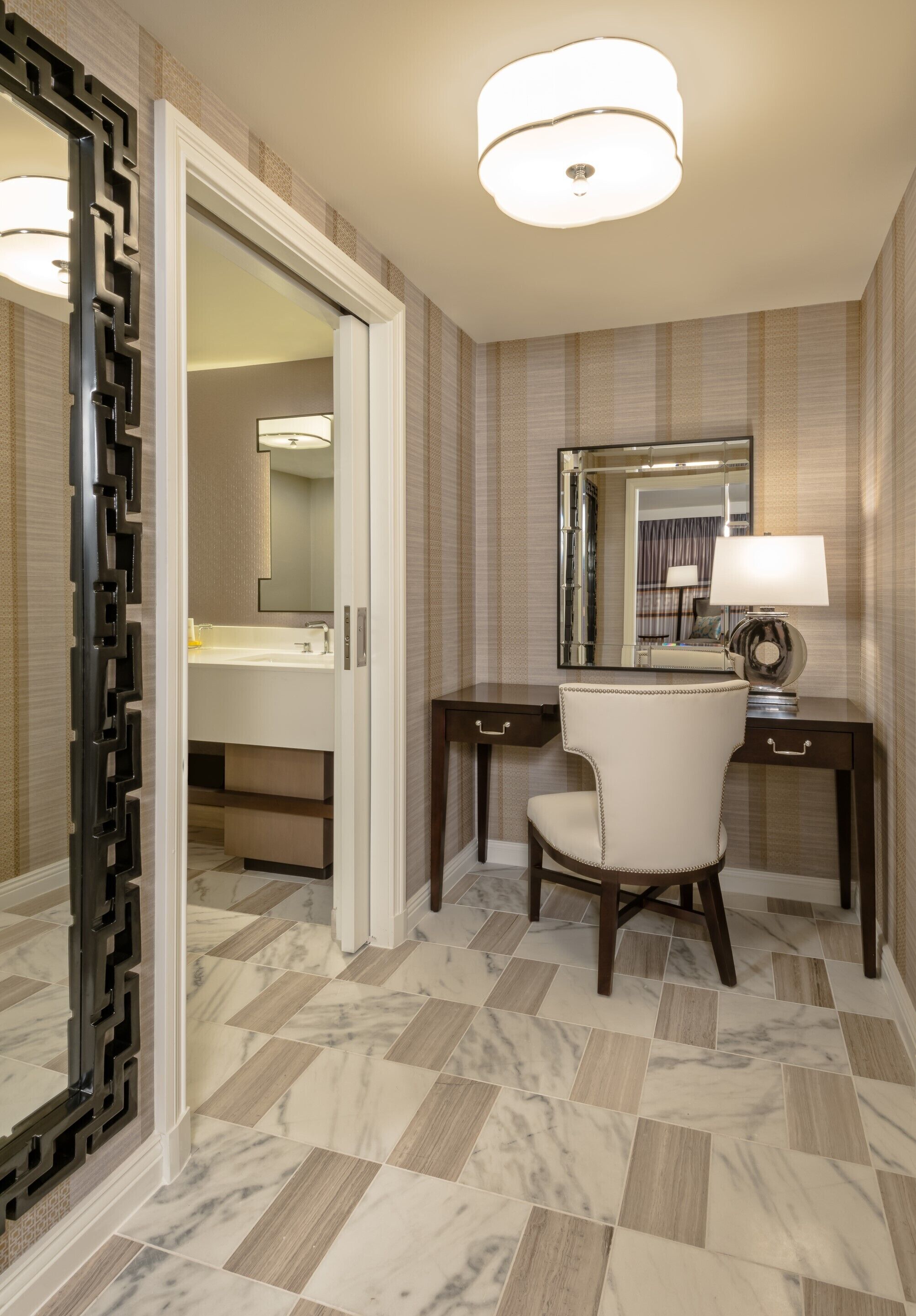 A modern bathroom with a marble floor, a large mirror, a desk with a chair, and a decorative wall panel.