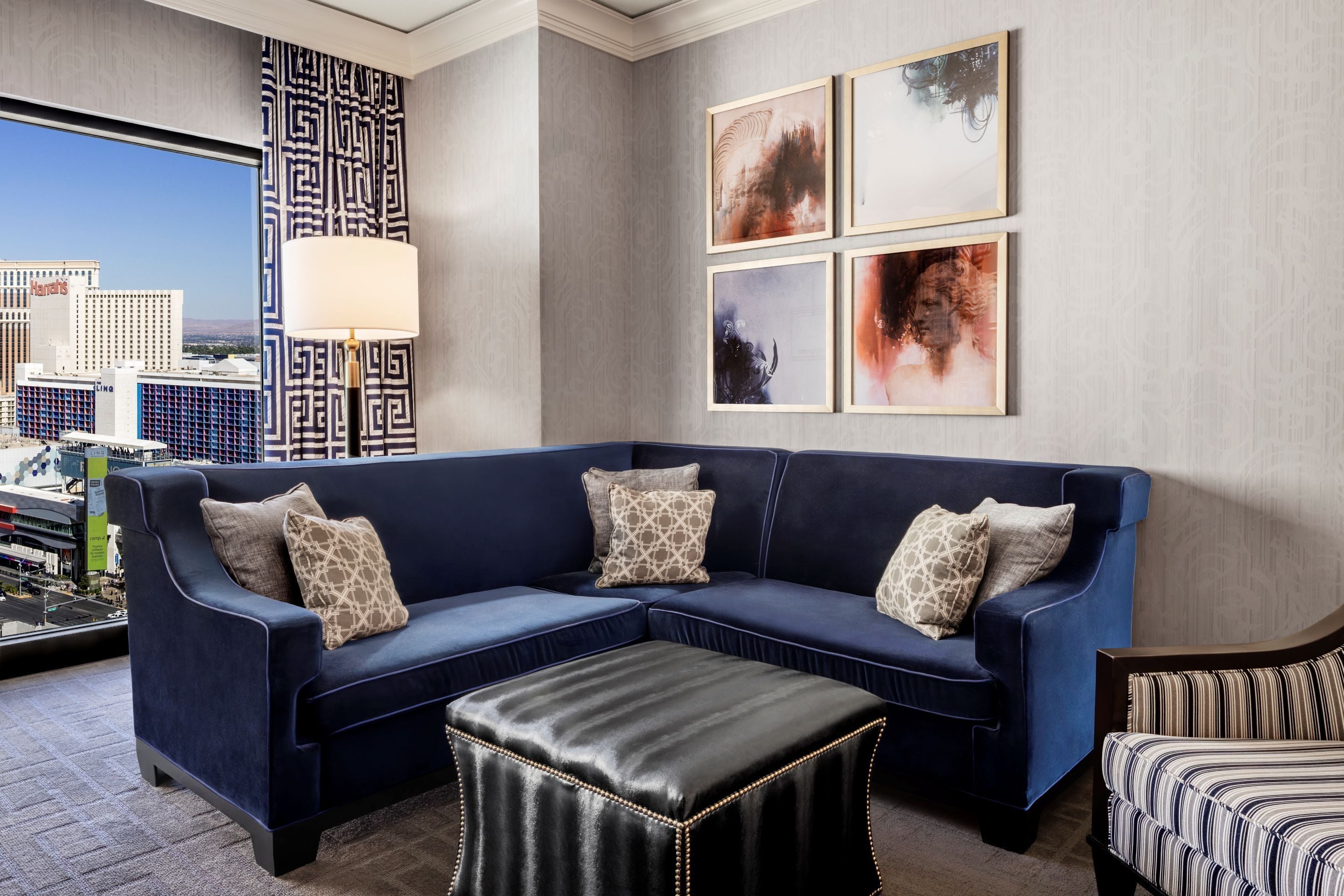 Een moderne woonkamer met een blauwe hoekbank, een leren poef en uitzicht op een stadsbeeld door een groot raam.