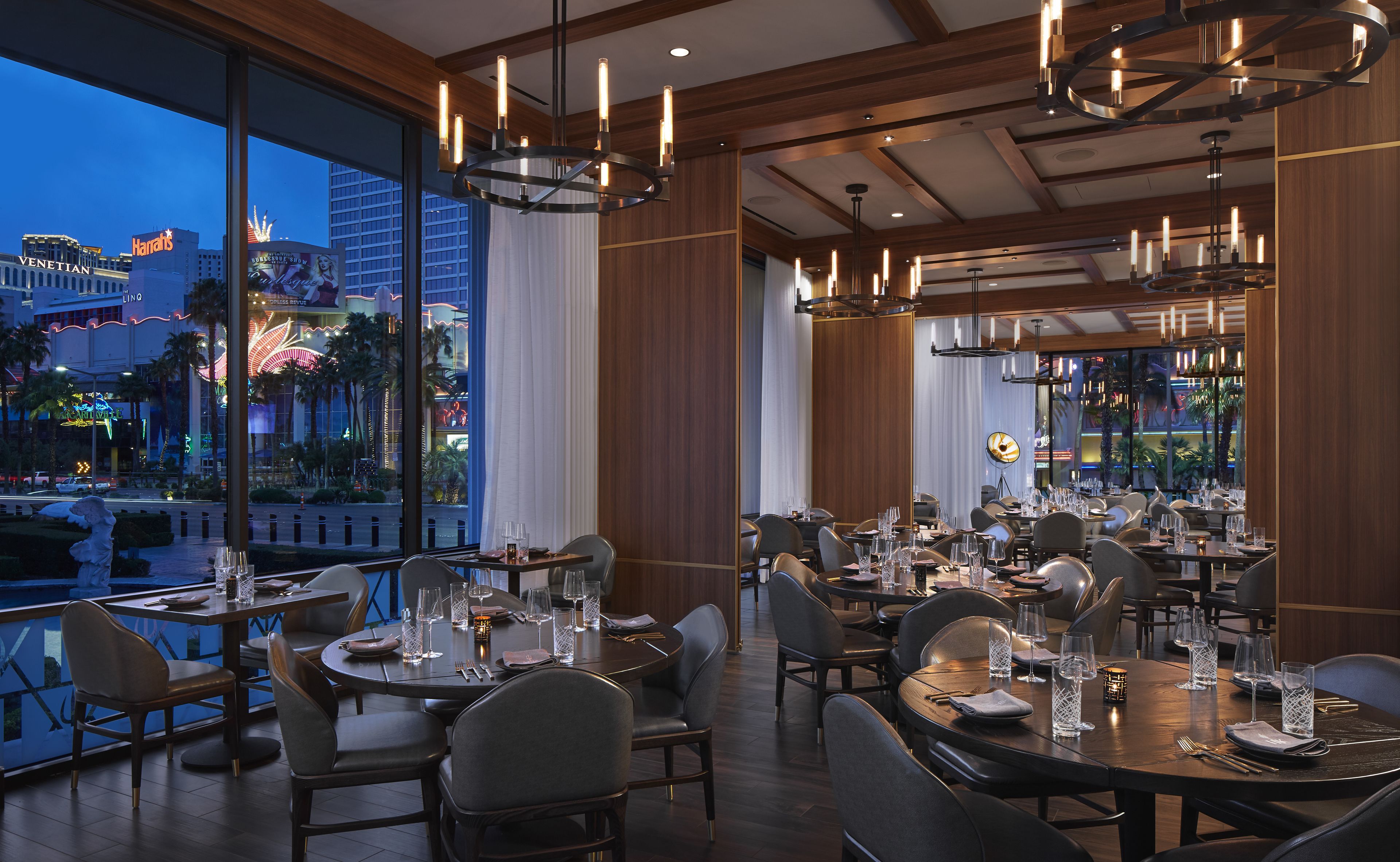 A restaurant with multiple dining tables set for guests, large windows offering a view of the cityscape, and modern lighting fixtures.