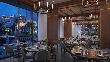 A restaurant with multiple dining tables set for guests, large windows offering a view of the cityscape, and modern lighting fixtures.