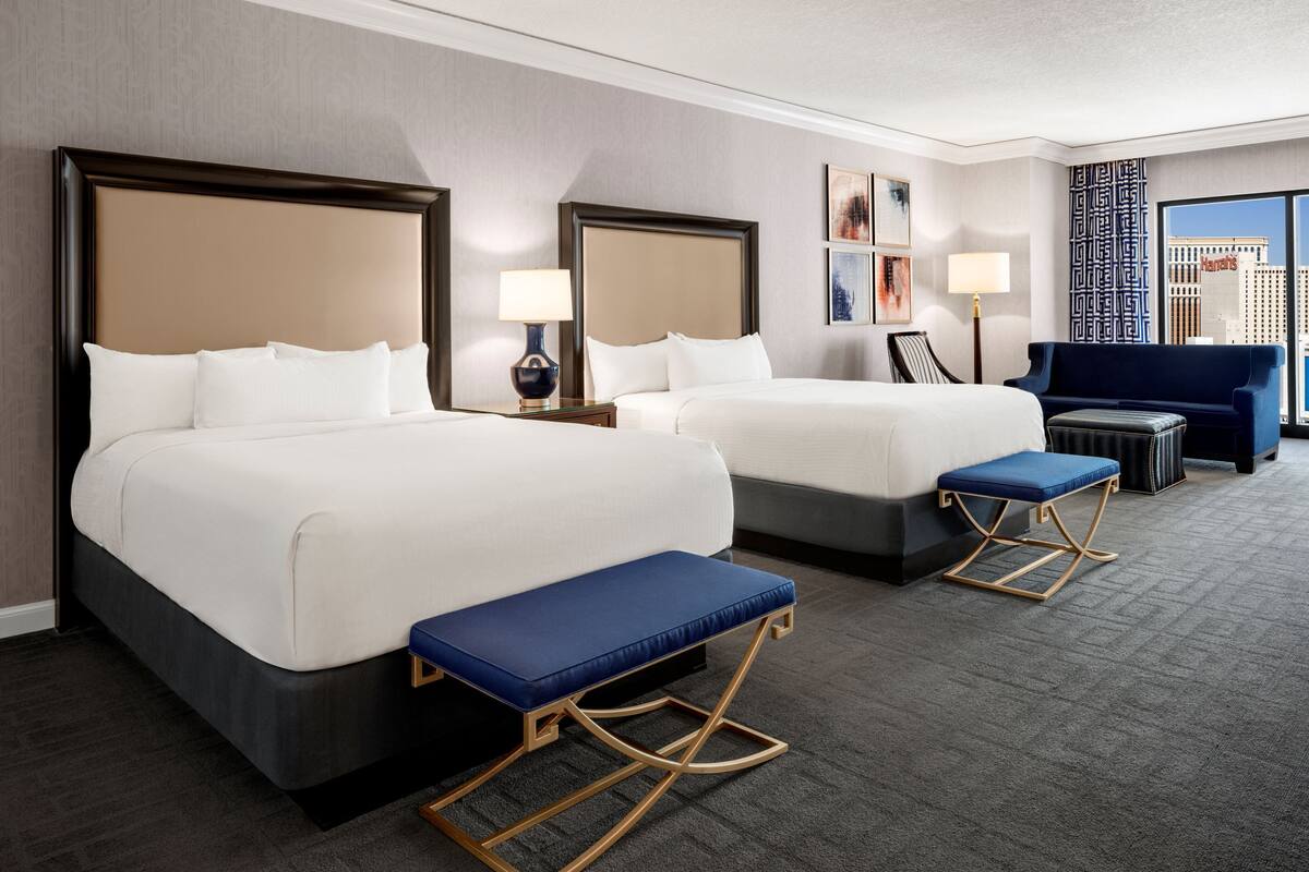 Une chambre d’hôtel avec deux lits, un banc bleu, un canapé et une grande fenêtre donnant sur la ville.