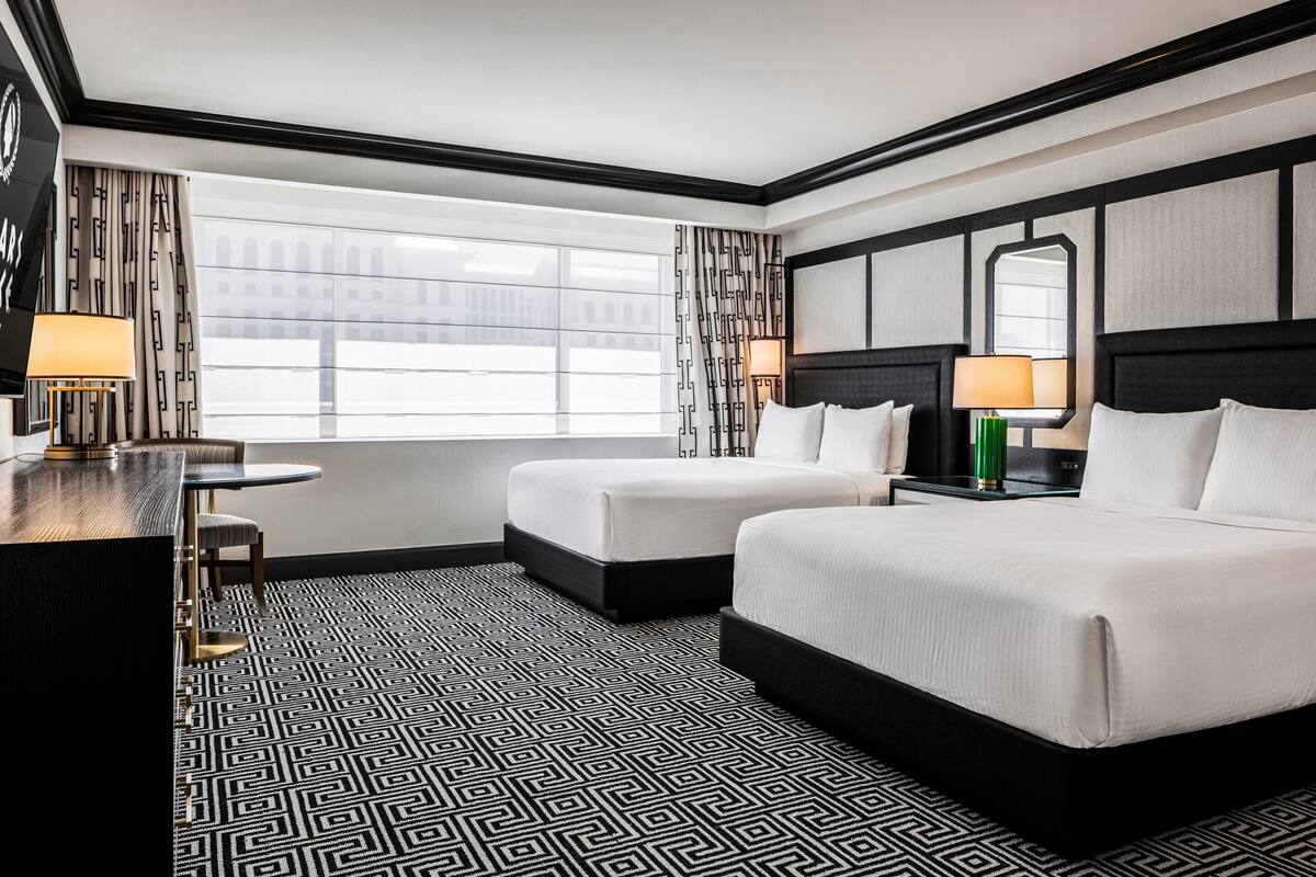Une chambre d’hôtel avec un grand lit, un lit plus petit, un bureau avec une chaise et une fenêtre avec des rideaux.