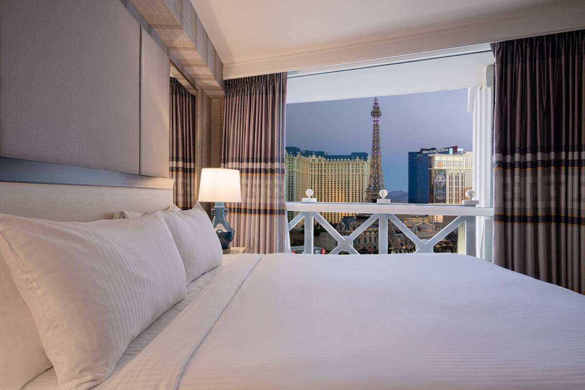 Une chambre d’hôtel avec un grand lit, une table de chevet avec une lampe et un balcon offrant une vue sur la Tour Eiffel et d’autres bâtiments.