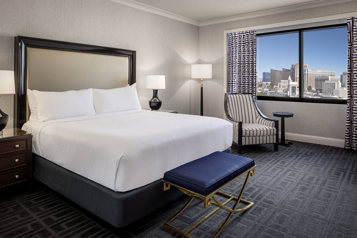 Une chambre d’hôtel avec un grand lit, une chaise, une petite table et une vue sur le paysage urbain par la fenêtre.
