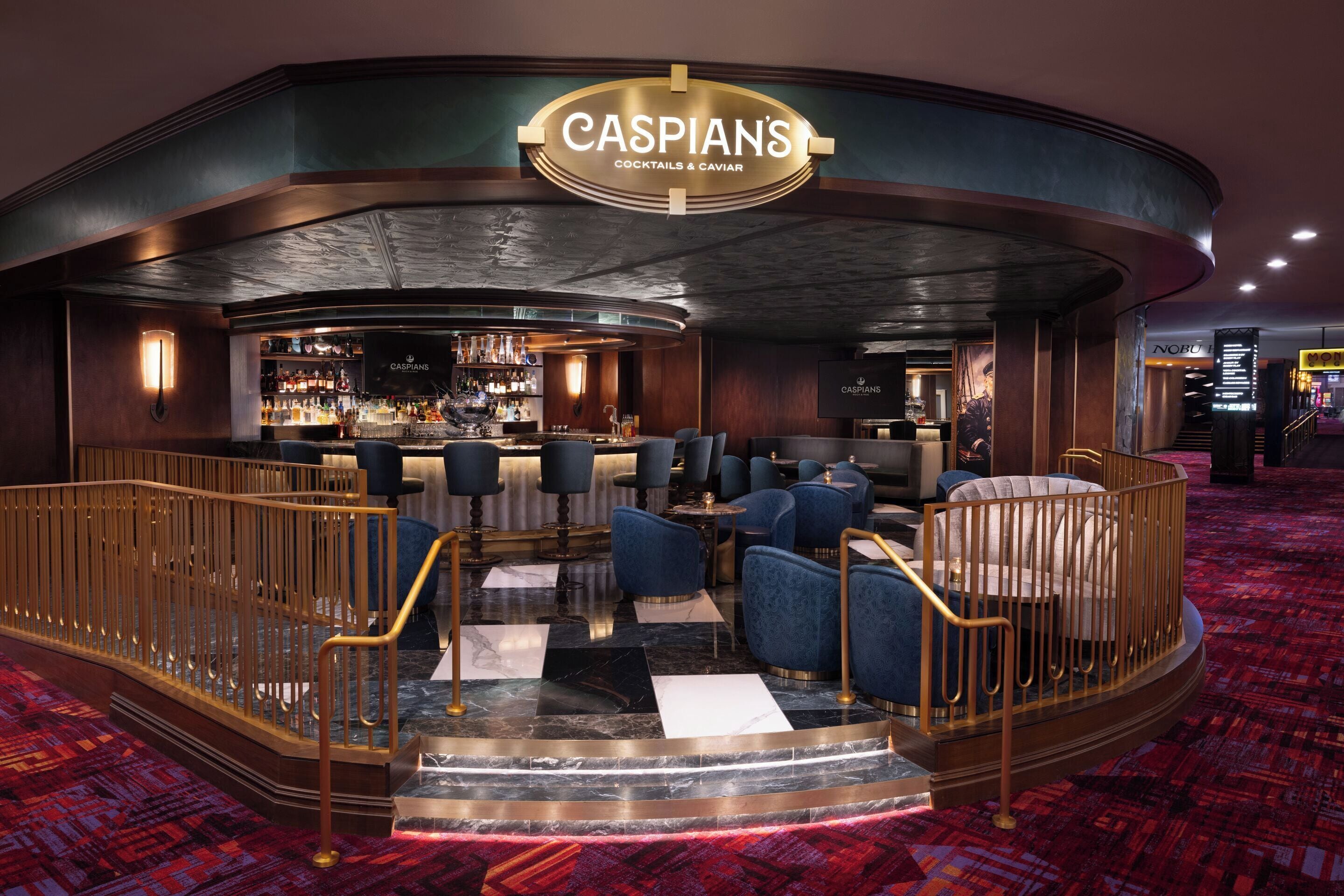 En bardel med en skylt som visar texten ”CASPIANS COCKTAILS & CAVIAR”, en bardisk, sittplatser med blå klädda stolar och en mönstrad matta.