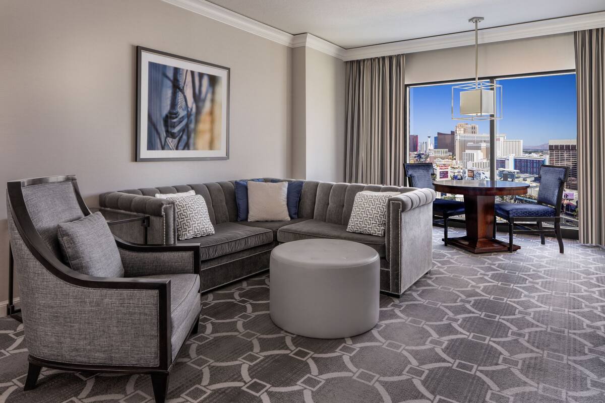 Une chambre d’hôtel moderne avec un canapé d’angle, un pouf rond, une table à manger avec des chaises, une grande fenêtre donnant sur la ville et une œuvre d’art encadrée au mur.