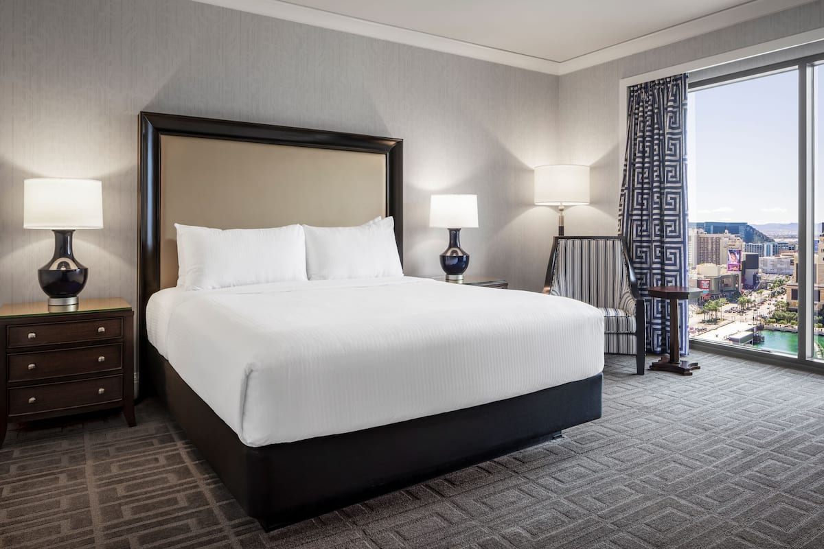 Une chambre d’hôtel avec un grand lit, deux tables de chevet avec des lampes, une vue sur le paysage urbain par une grande fenêtre et une moquette à motifs.