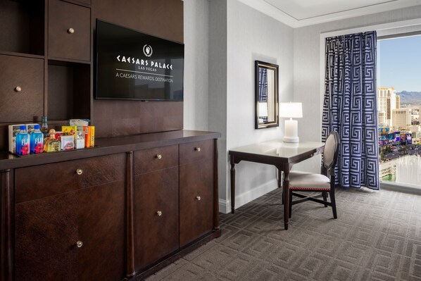 A hotel room with a dark wood cabinet, a flat-screen TV, a desk with a chair, and a view of the city.