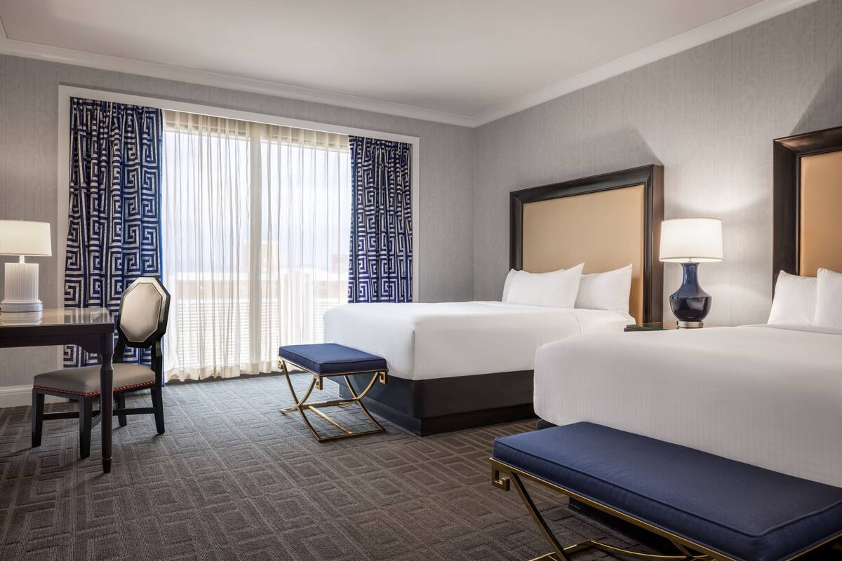 Une chambre d’hôtel avec deux lits, un bureau, une chaise et une fenêtre avec des rideaux à motifs.