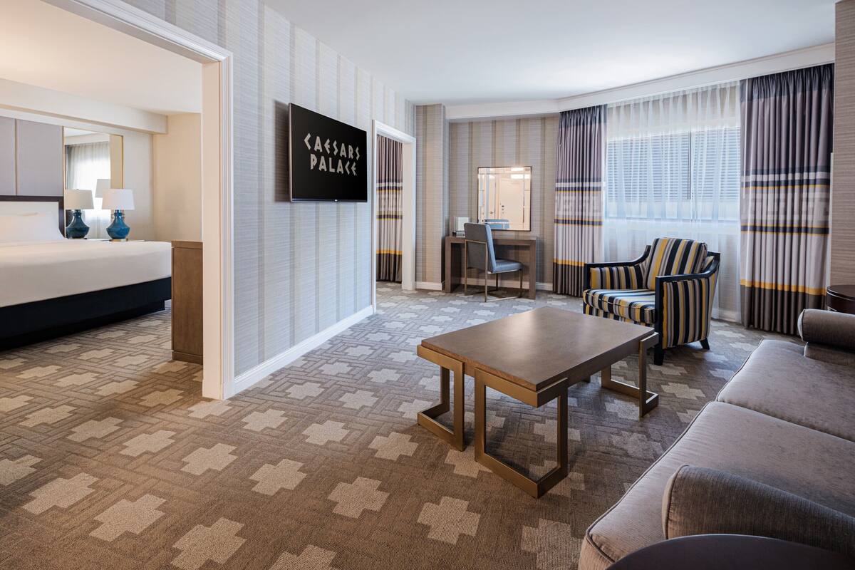 Une chambre d’hôtel équipée d’un lit, d’un bureau, d’une chaise, d’un canapé et d’un écran de télévision affichant l’image de « Caesars Palace ».