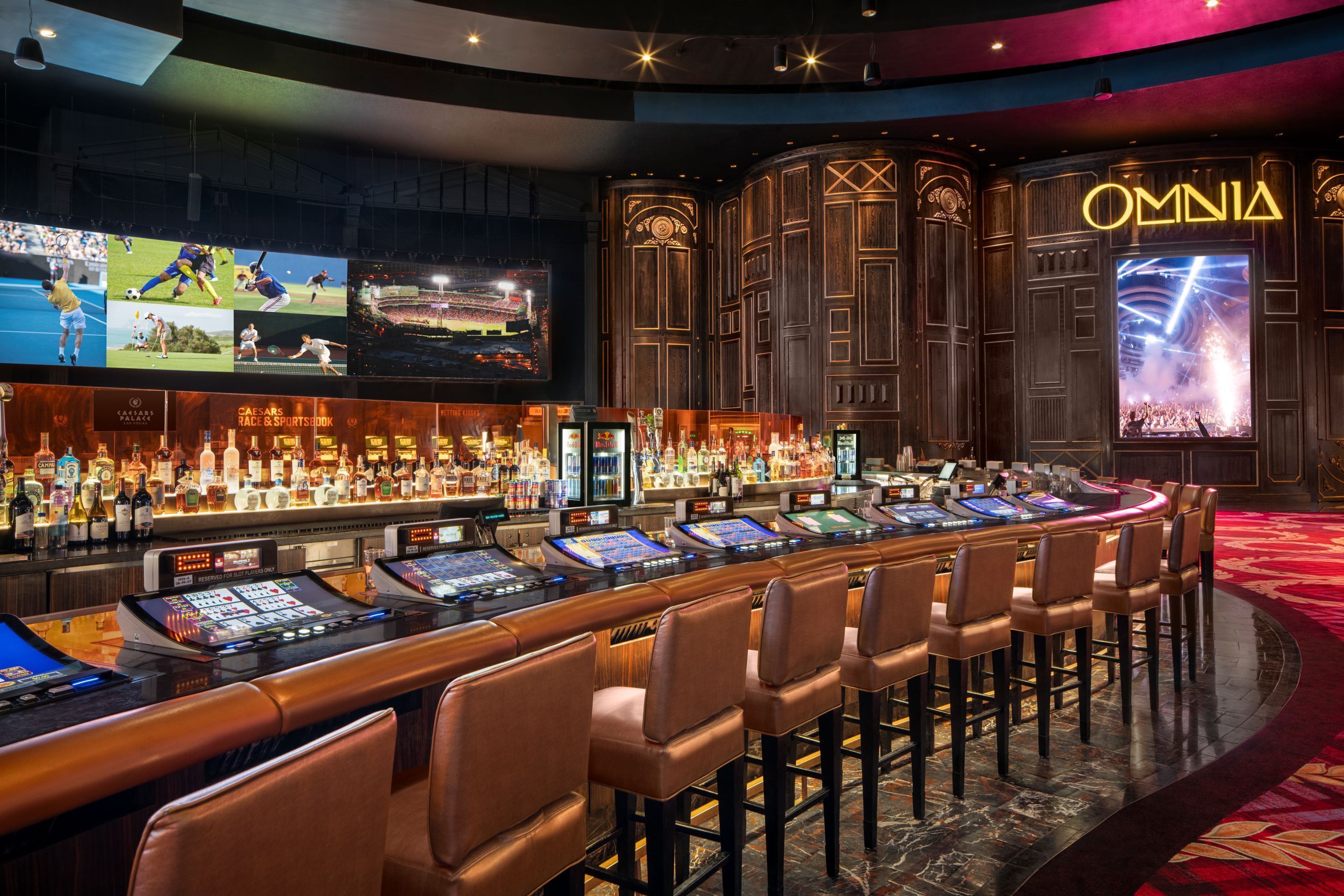 A bar area with multiple gaming machines, bar stools, and a large screen displaying sports events.