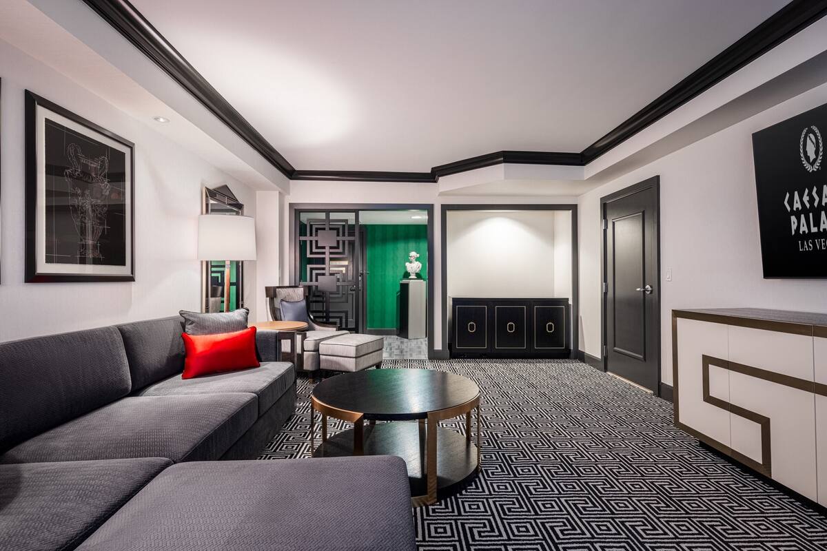 Un hall d’hôtel moderne avec un canapé gris, une table basse ronde et un sol à motifs. On aperçoit un écran vert à l’arrière-plan, et un panneau est accroché au mur.