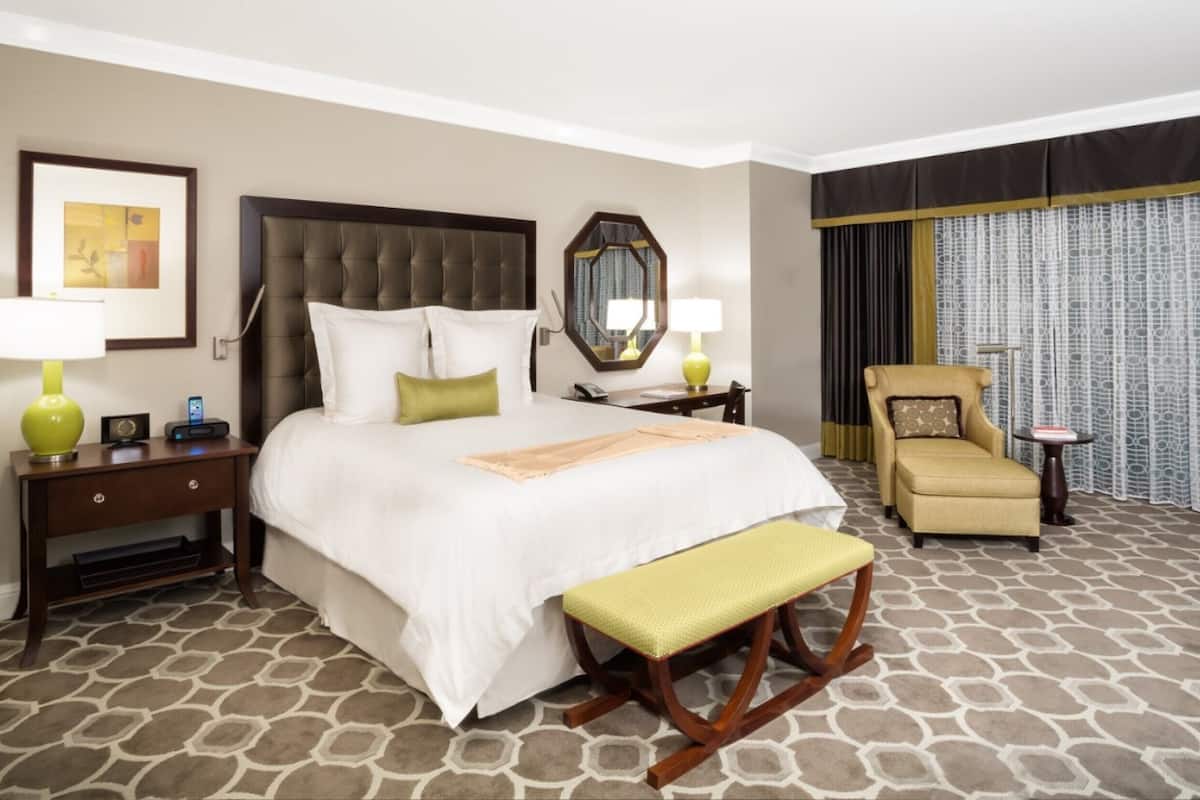 Une chambre d’hôtel avec un grand lit, une table de chevet, une chaise, un banc, un miroir et une fenêtre avec des rideaux.