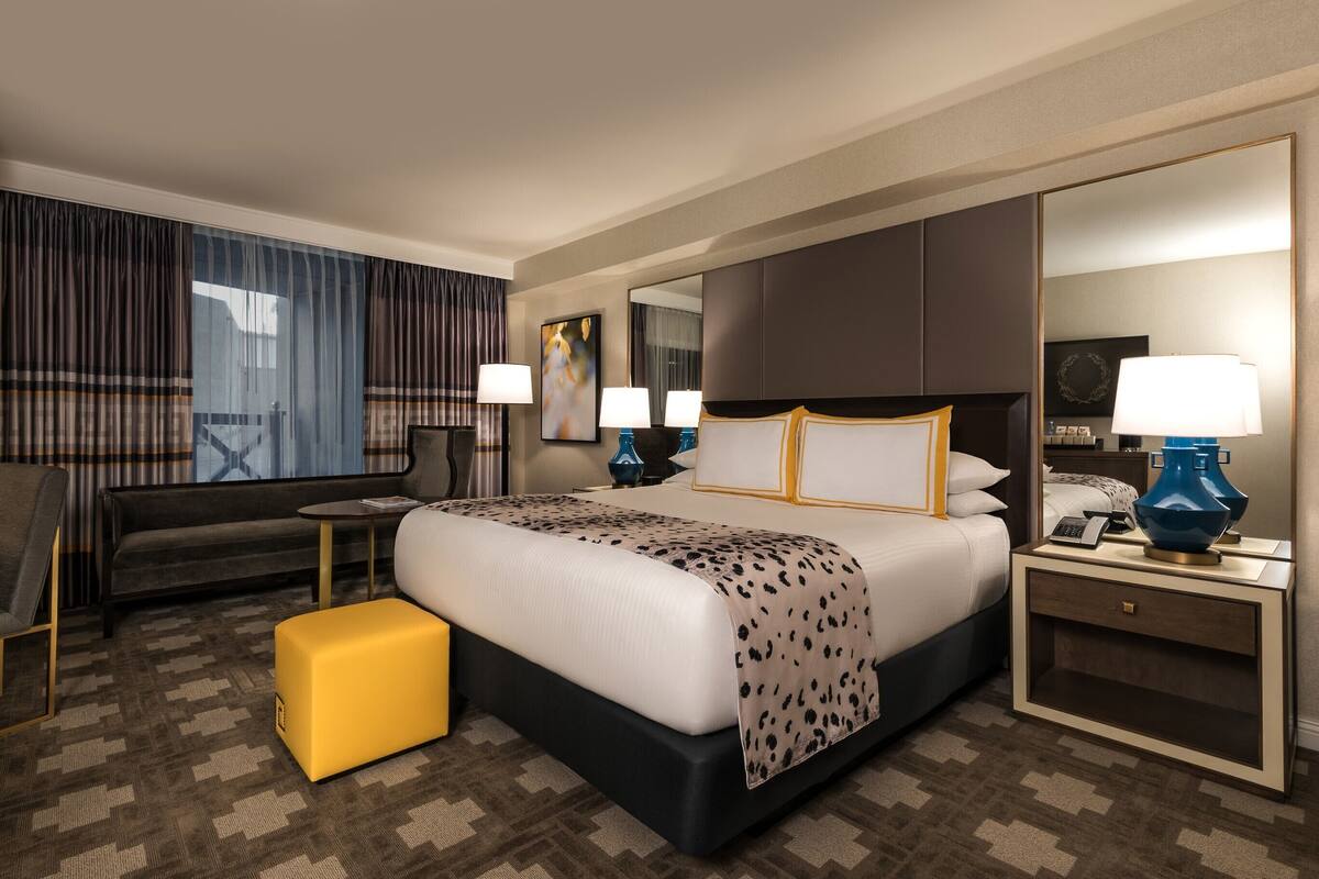 Une chambre d’hôtel avec un grand lit, un canapé, un pouf jaune, un bureau avec une lampe et un grand miroir.