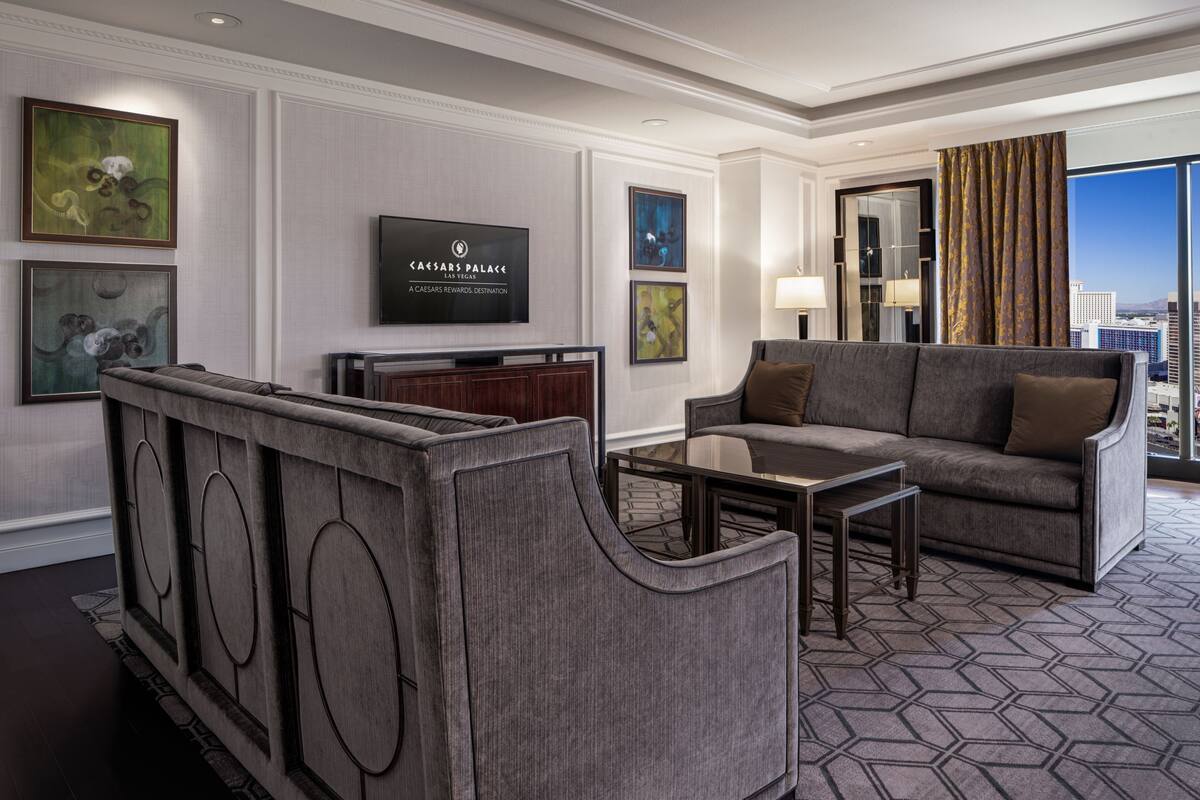 Un hall d’hôtel moderne avec un canapé gris, une petite table, un téléviseur fixé au mur et des œuvres d’art encadrées.