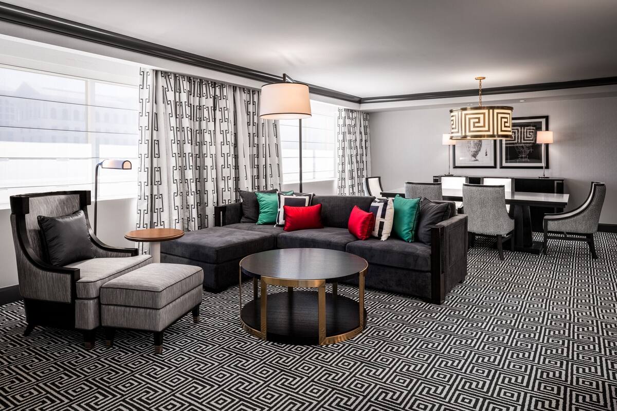 Une chambre d’hôtel moderne comprenant un canapé d’angle, des fauteuils, une table basse ronde et un coin repas avec des chaises et une table.