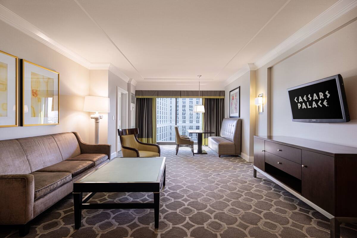 Une chambre d’hôtel équipée d’un canapé, d’une table basse, d’une télévision et offrant une vue sur la ville grâce à de grandes fenêtres.