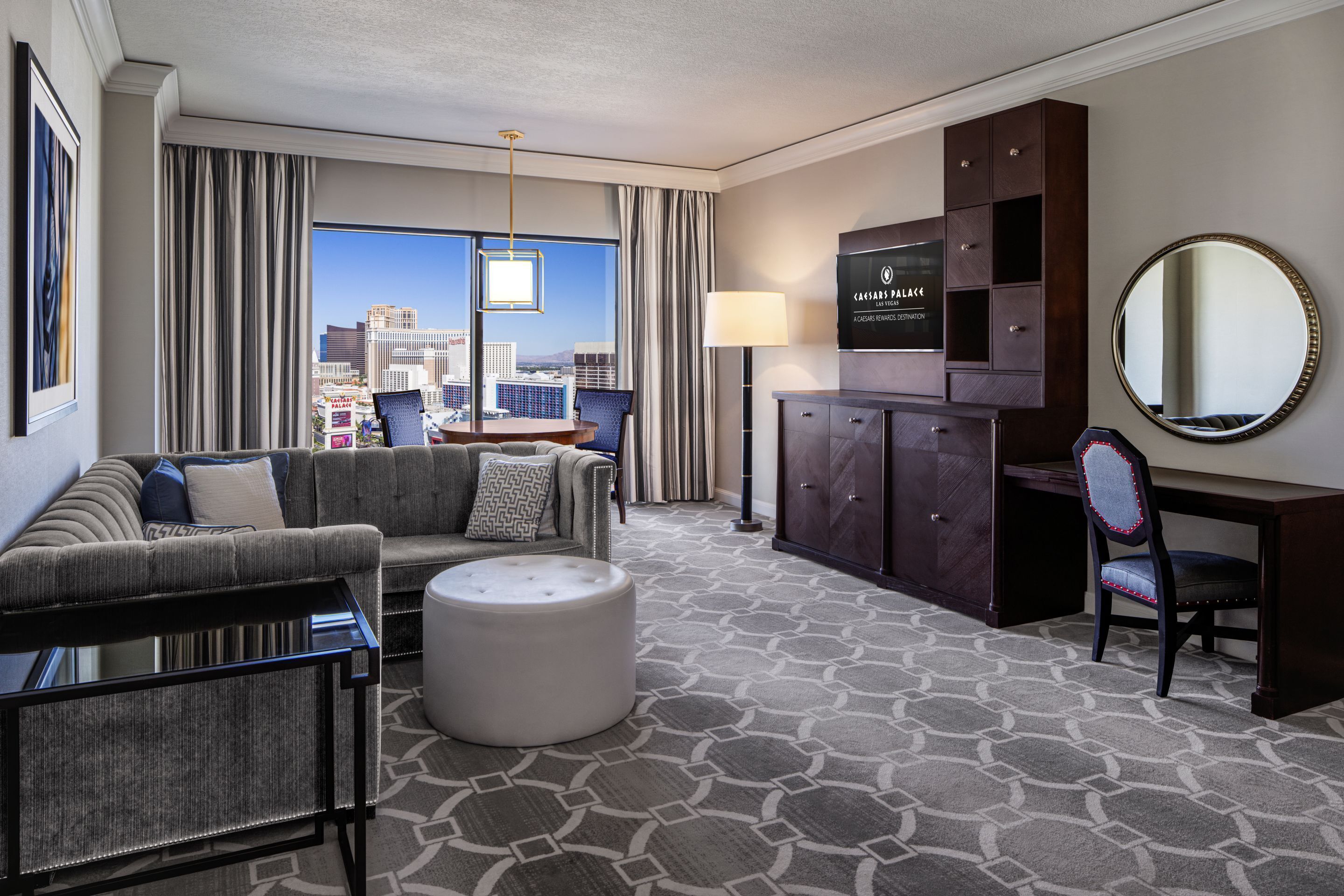 A hotel room with a city view, a sofa, a desk, a chair, a TV, and a lamp.