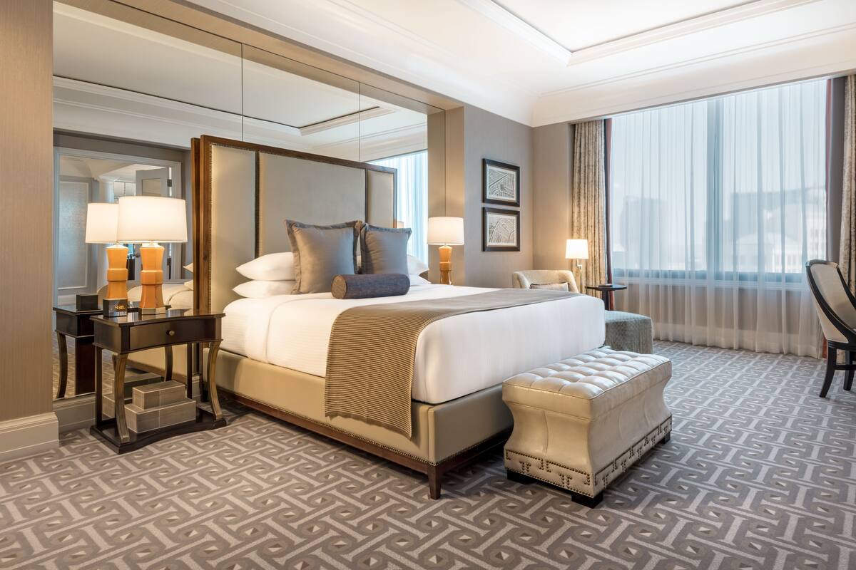 Une chambre d’hôtel avec un grand lit, un coin salon avec un fauteuil, un bureau et une grande fenêtre avec des rideaux.