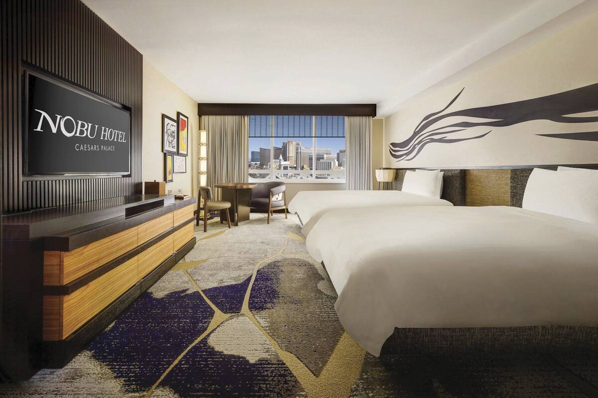 Une chambre d’hôtel avec un grand lit, un bureau, un téléviseur diffusant des images de l’hôtel « Nobu Hotel Caesars Palace », et une vue sur le paysage urbain.