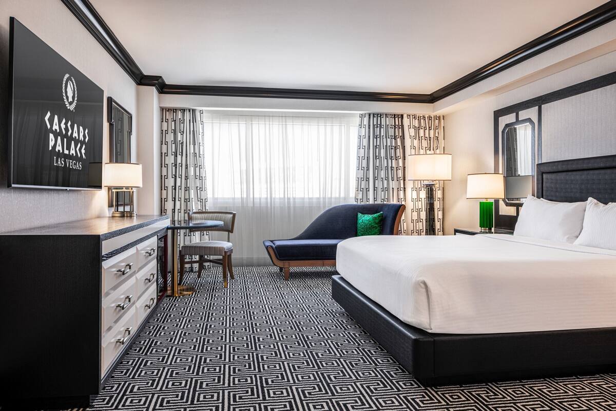 Une chambre d’hôtel avec un grand lit, un bureau avec des tiroirs, une chaise, une fenêtre avec des rideaux et un téléviseur fixé au mur.