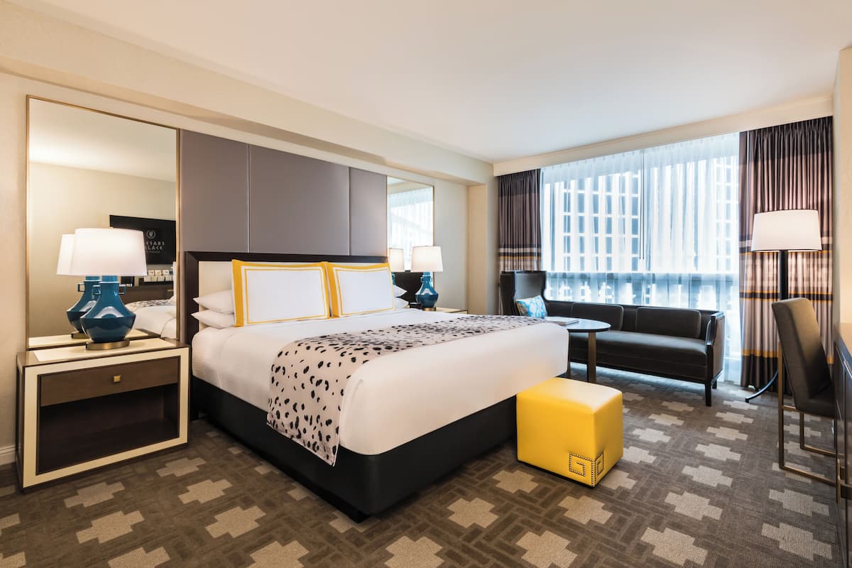 Une chambre d’hôtel avec un grand lit, des tables de chevet, un canapé, une petite table et une grande fenêtre avec des rideaux.