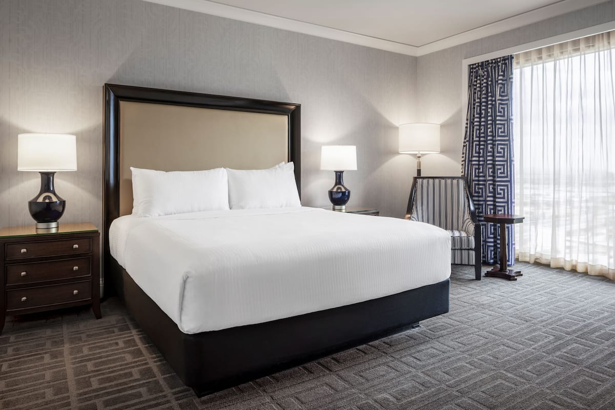 Une chambre d’hôtel avec un grand lit, deux tables de chevet avec des lampes, une chaise et une fenêtre avec des rideaux.