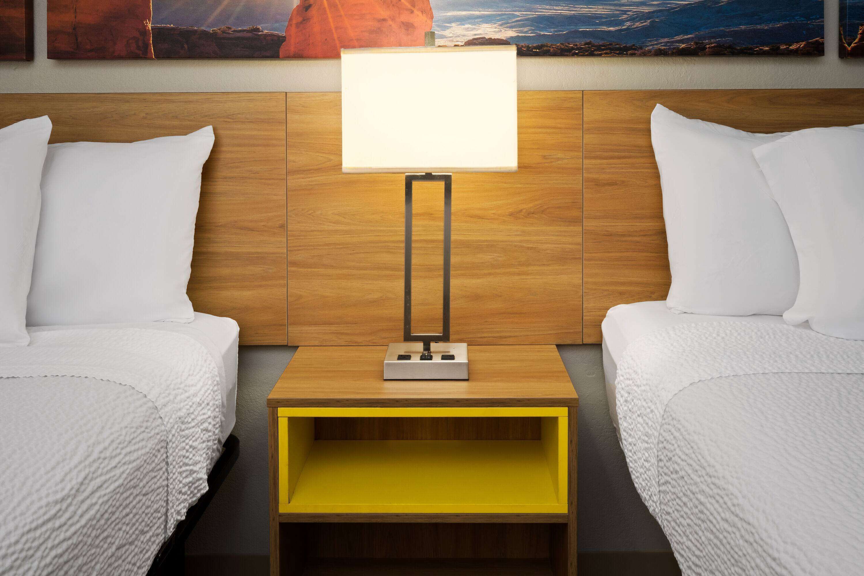 Quarto de hotel com duas camas, cabeceira de madeira, mesa de cabeceira com abajur e uma estante amarela.