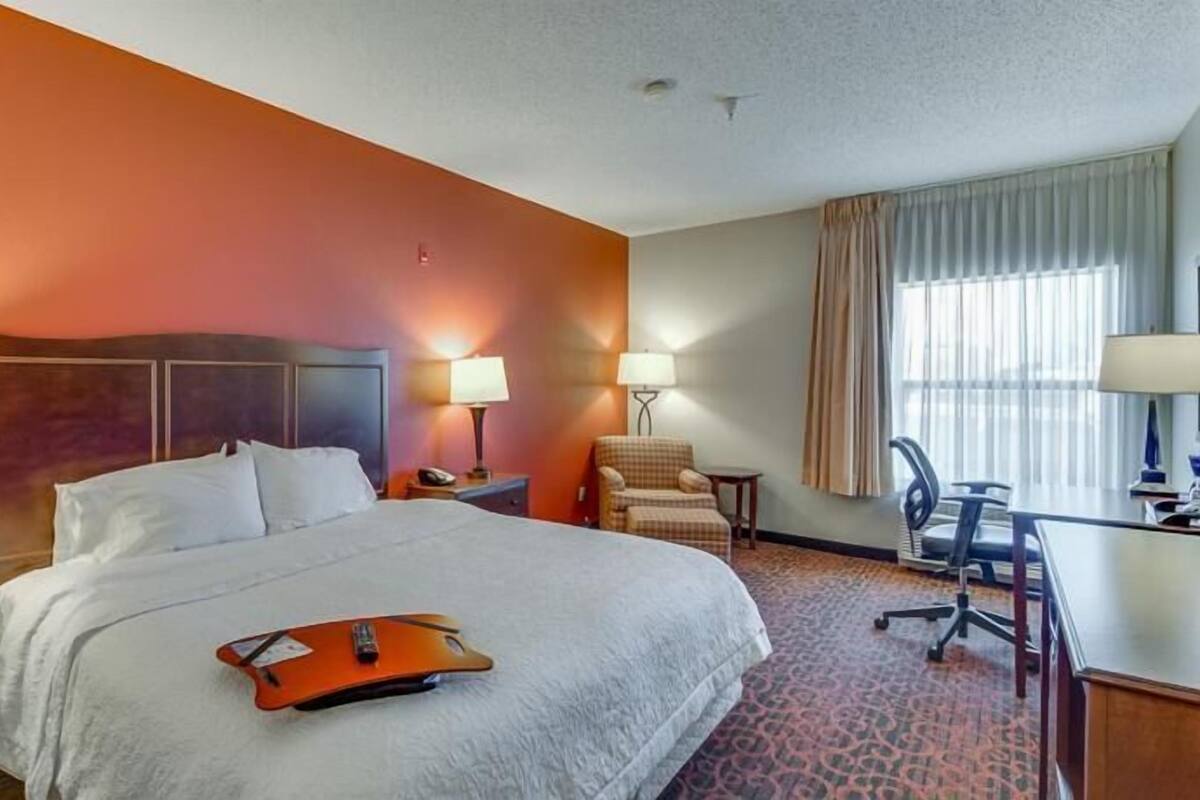 Une chambre d’hôtel comprenant un lit, deux tables de chevet avec des lampes, un bureau avec une chaise et une grande fenêtre avec des rideaux.