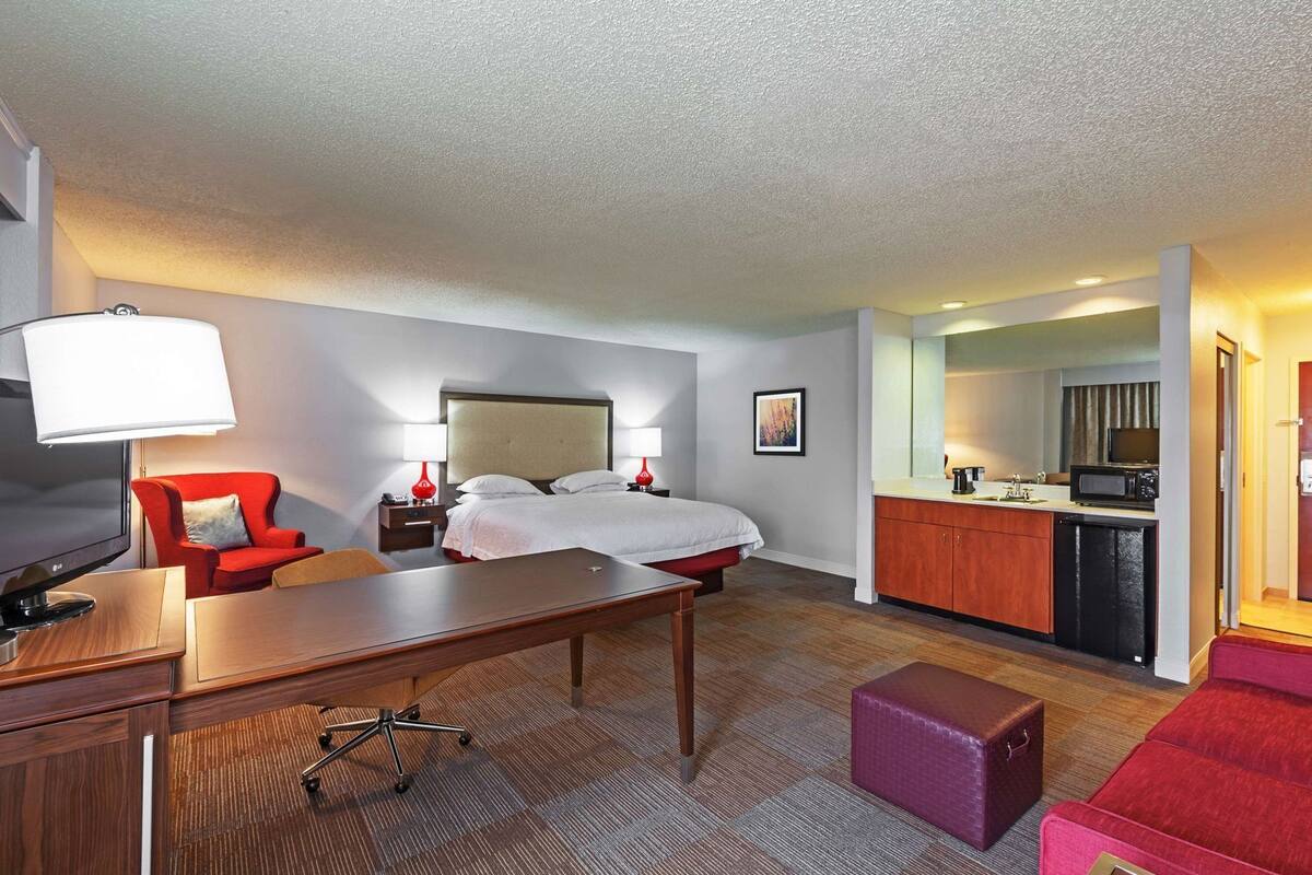 A hotel room with a bed, desk, chair, kitchenette, and a red armchair.
