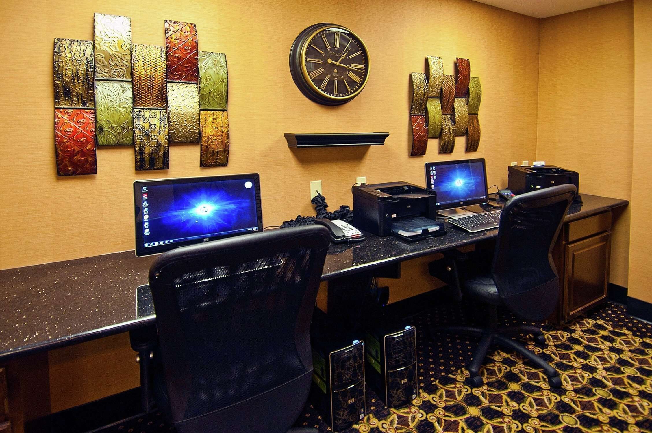 Habitación de hotel con un escritorio grande, dos sillas, dos computadoras de escritorio, un reloj y una obra de arte colgada en la pared.