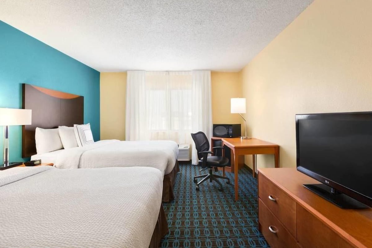 A hotel room with two beds, a desk with a TV, a chair, and a lamp.