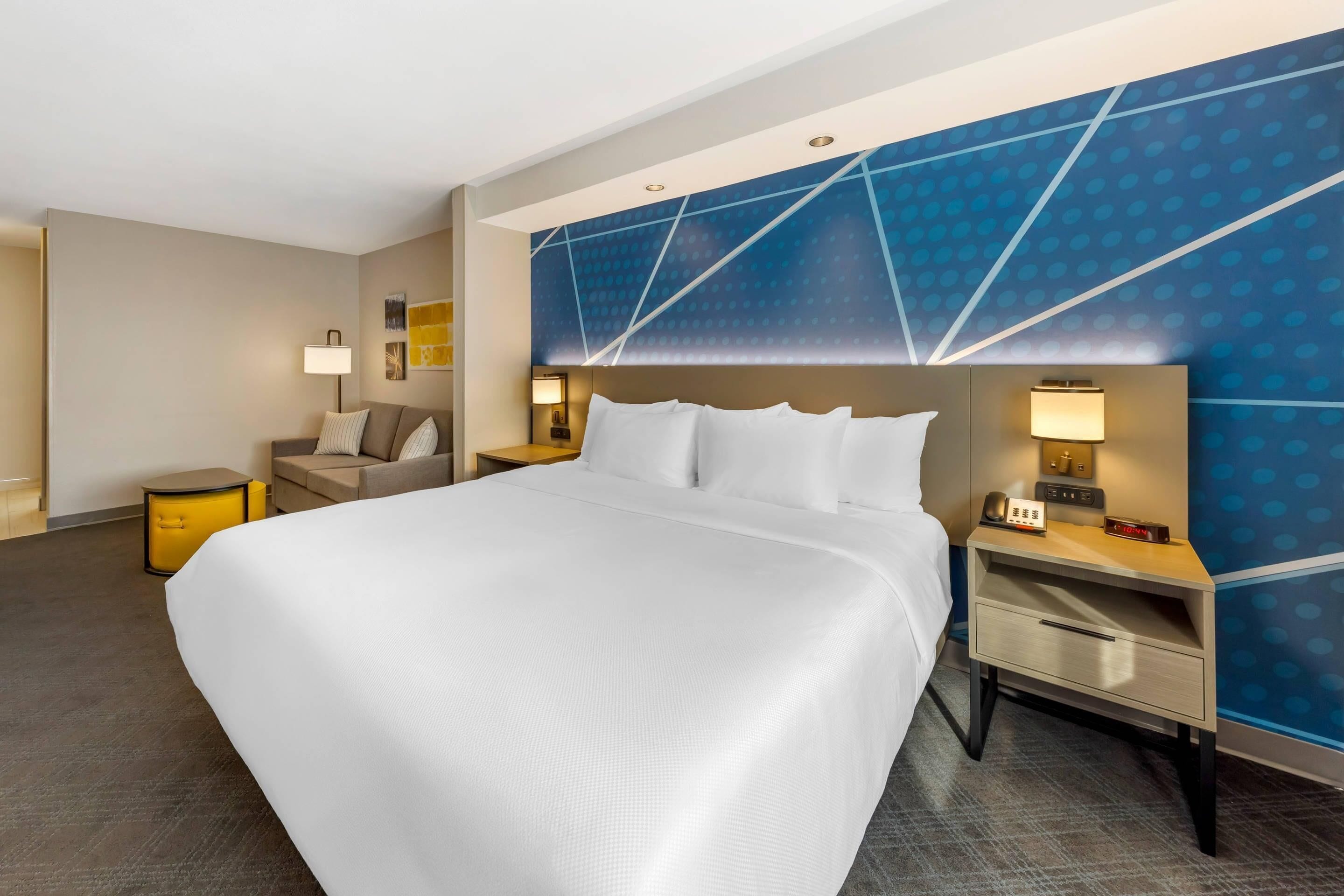 A hotel room with a large bed, a bedside table with a lamp, a small side table, and a wall with a geometric pattern.