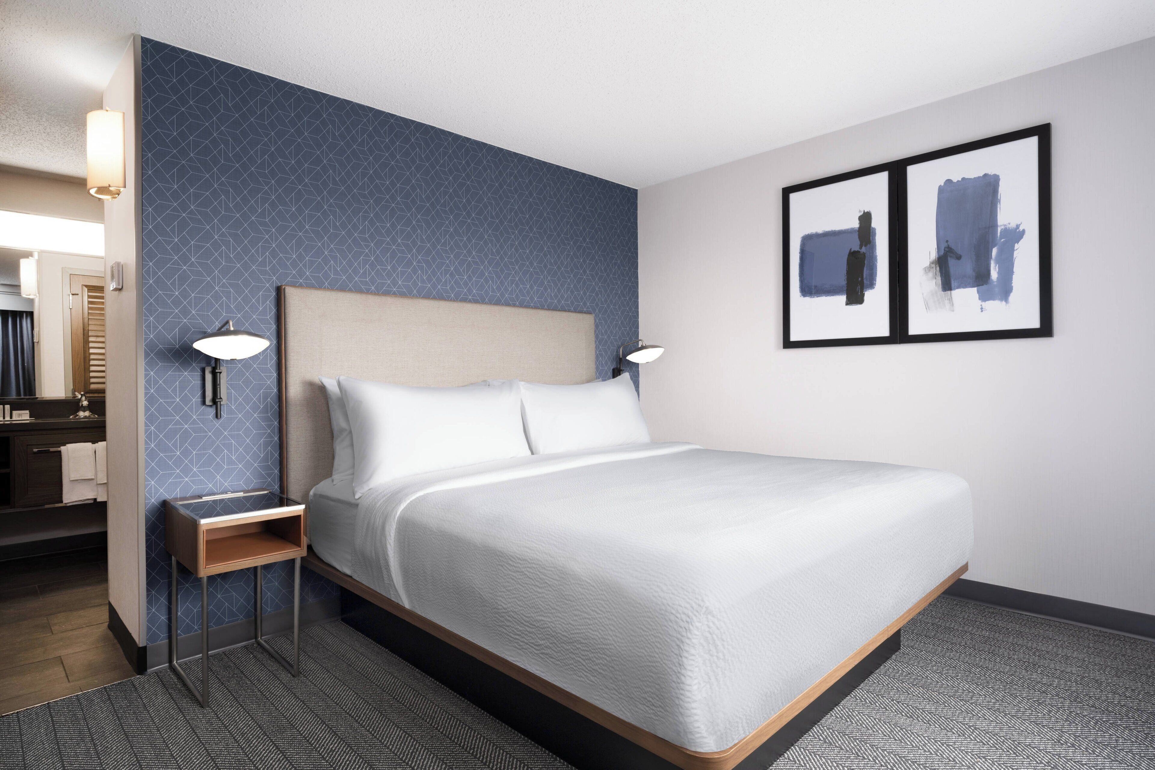 Una habitación de hotel con cama, mesitas de noche, un baño con espejo y dos cuadros abstractos enmarcados en la pared.