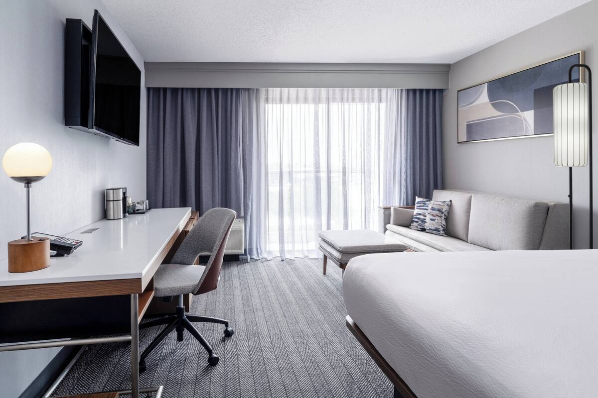 A hotel room with a bed, a desk with a lamp, a chair, a TV mounted on the wall, and a large window with curtains.