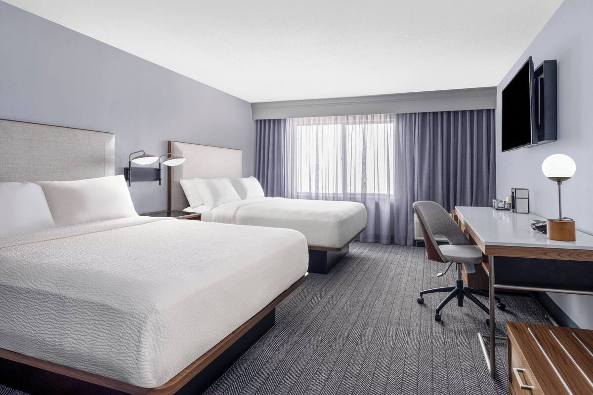 A hotel room with two beds, a desk, a chair, a TV, and a window with curtains.
